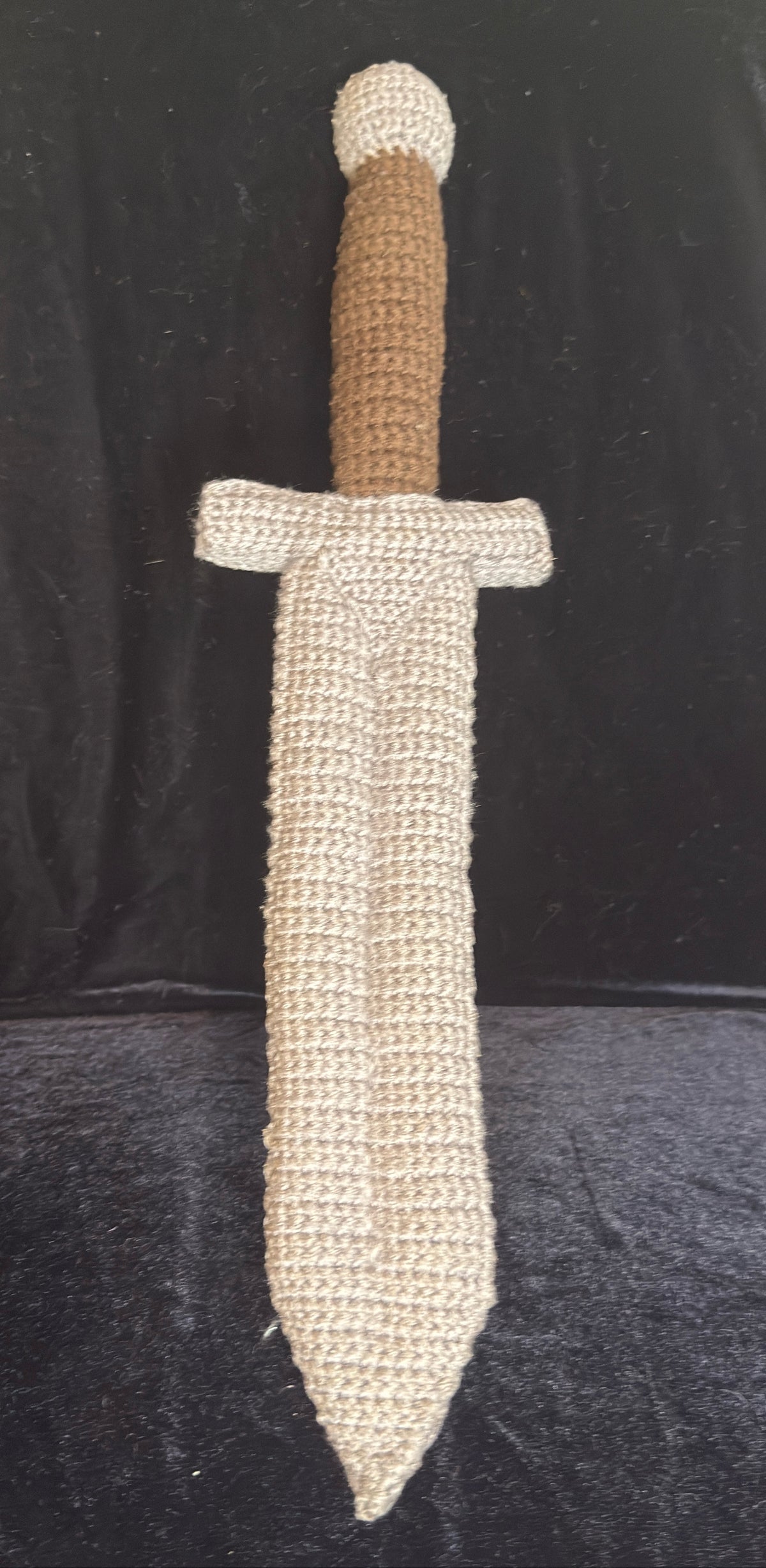 A crocheted toy or decorative sword with a textured gray blade and guard, and a brown handle, displayed against a black fabric background.