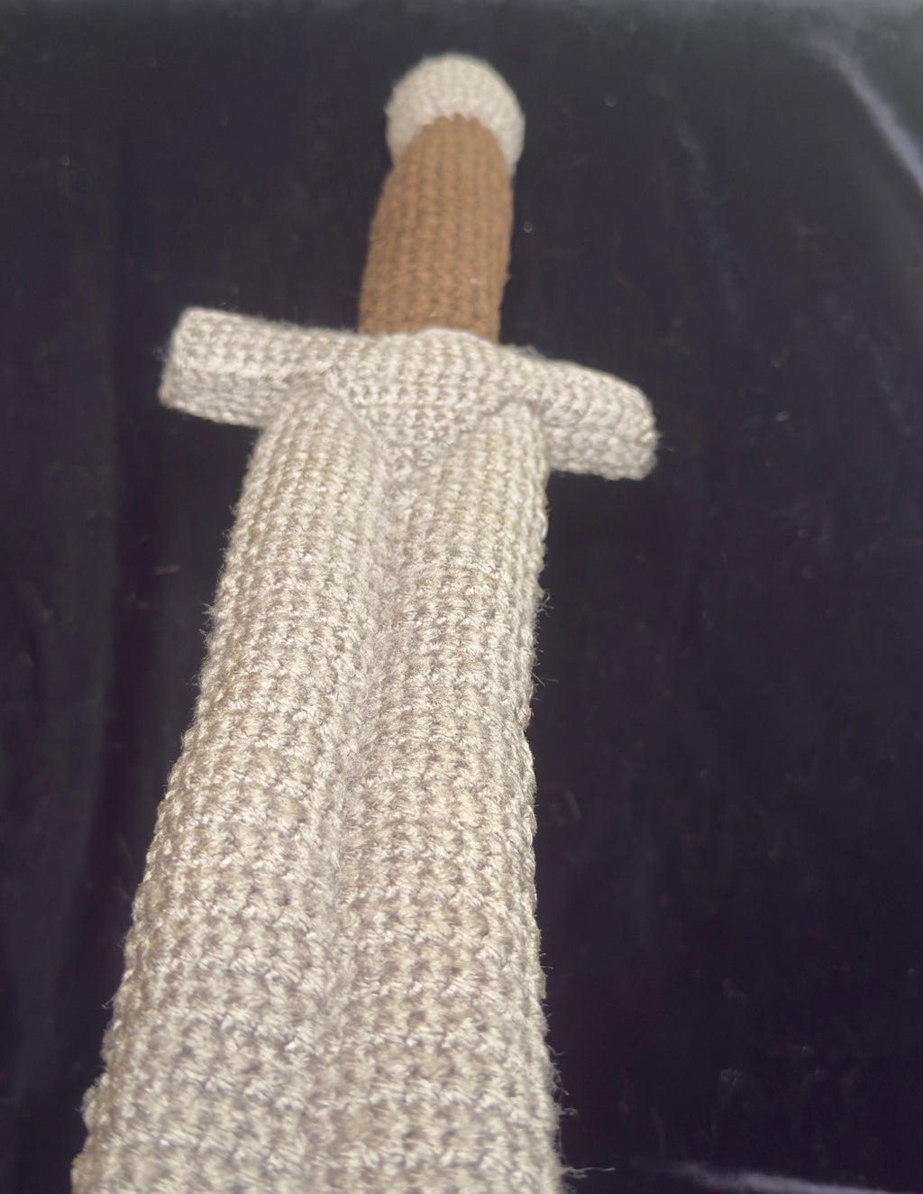 A close-up of a crocheted sword cover in a textured gray yarn, with a brown handle and gray pommel, shown against a dark black background.